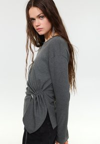 Siv dolg rokav z okroglim izrezom, naborki v pasu in večplastnim zadnjim dizajnom. Mehka tekstura in sproščen kroj.