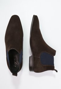 Bottes Chelsea en daim marron avec panneaux latéraux élastiques bleus et bouts arrondis. Semelle en caoutchouc plate avec un léger talon. Finition supérieure texturée.