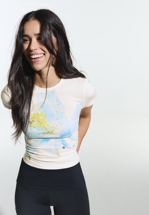 Young woman with long dark hair smiling, wearing a white t-shirt with a large blue and yellow flower print and black pants.