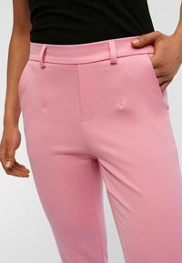 Pantalons tailleur roses avec une texture lisse, dotés d'une taille plate, de poches latérales et d'une coupe ajustée.