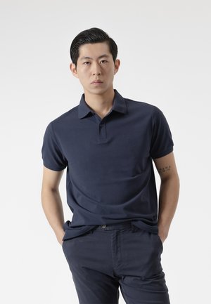 Joven con las manos en los bolsillos, vestido con una camisa polo azul marino oscuro y pantalones a juego, de pie contra un fondo claro y liso.