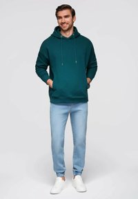 Sudadera con capucha en color verde azulado con cordones, combinada con jeans jogger de color azul claro. Zapatillas deportivas blancas completan el conjunto. Textura plana y suave en toda la prenda.