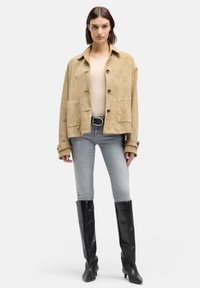 Kurtka z khaki zamszu o luźnym kroju, z dwoma przednimi kieszeniami i czarnym zapięciem na guziki, zestawiona z jasnoniebieskimi dżinsami i czarnymi wysokimi kozakami za kolano.
