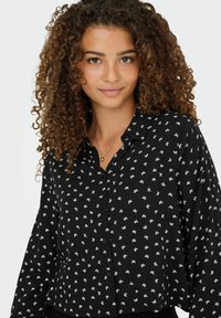 Jeune femme aux cheveux bouclés portant une chemise noire à boutons avec de petits motifs de nœuds blancs et un collier en or, souriant doucement.