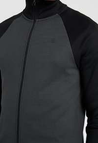 Giacca a zip grigio scuro con maniche raglan nere, tessuto strutturato, colletto alto e un piccolo logo sul petto. Finitura liscia nel complesso.