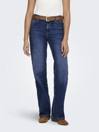 Femeie purtând blugi bleumarin cu croială bootcut, curea maro, top alb și pantofi maro din piele întoarsă, stând în picioare pe un fundal simplu și deschis la culoare.