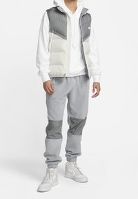 Chaleco de plumas en gris y crema con cierre frontal, combinado con pantalones de forro polar grises que tienen paneles grises, y zapatillas altas blancas.