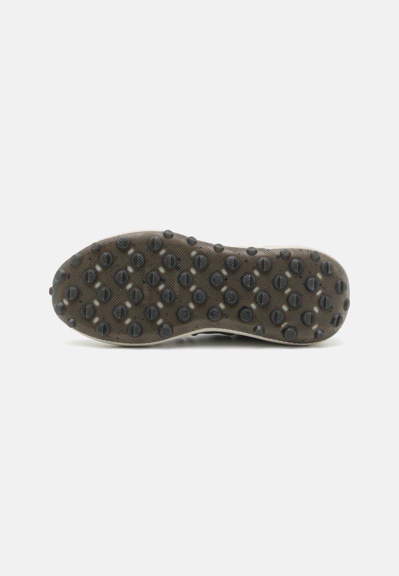 La semelle de la chaussure de sport présente une surface en caoutchouc noir texturé avec de petites grips circulaires, délimitée par une bordure beige contrastante.