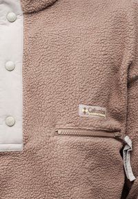 Μπεζ φλις μπουφάν Columbia Sportswear με τσέπη με φερμουάρ και κουμπιά με πάτημα, που δείχνει υφή απαλού υφάσματος και μερική ετικέτα με λογότυπο.