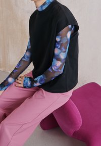 Černý top s krátkým rukávem, pod ním květinová košile s dlouhým rukávem, nošena s růžovými kalhotami širokého střihu, sedící na růžovém nepravidelném polštáři.