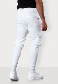 Weiße Jogginghosen aus Baumwollmischung, mit schmaler Passform, elastischem Bund, seitlicher Reißverschlusstasche und gestickten Details. Kombiniert mit weißen Sneakers.