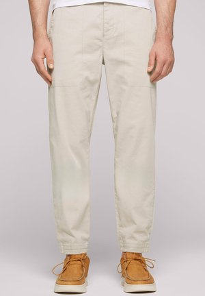 Pantalones cargo béis color beige, ajustados en los tobillos, con bolsillos laterales y detalles de costura, combinados con unos zapatos de ante marrones con cordones.