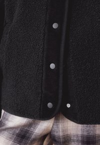 Veste polaire noire à boutons, présentant une surface texturée et des accents en tissu lisse, associée à un pantalon à carreaux à motif.