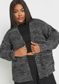 Kofta gjord av en strukturerad svart och grå stickning. Öppen fram, långa ärmar och avslappnad passform, bärs över en svart topp.