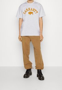 Grå kortærmet T-shirt med orange "Carhartt" tekst og bison grafik, parret med tan afslappede bukser og sorte snørresko.