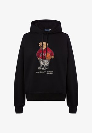 Zwarte capuchontrui met een geborduurde beer in een rode hoodie die een basketbal vasthoudt. Voorzien van witte sneakers en logodetails.