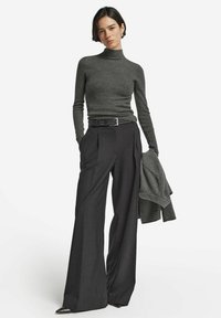 Dunkelgrauer, gerippter Rollkragenpullover kombiniert mit hochtaillierten, weiten anthrazitfarbenen Hosen und einem passenden Blazer, der über einem Arm hängt.