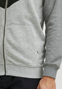 Gray zip-up hoodie made of soft cotton blend, featuring a black upper panel, ribbed cuffs, side pockets, and a small logo on the hem.