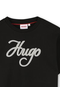 Crna majica kratkih rukava s okruglim izrezom koja na prednjoj strani ima veliki, kurzivnim, metalik srebrni logo "Hugo". Mekani teksturirani materijal.