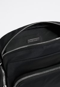 Interno di una borsa nera in nylon con numerosi scomparti con zip, texture liscia e una placca logo in metallo silver.