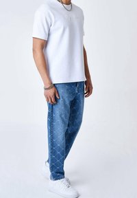Camiseta de algodón blanca con diseño en relieve, combinada con pantalones de mezclilla azul con estampado. Calzado: zapatillas blancas, pulsera de cadena de plata en la muñeca.