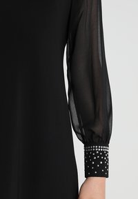 Manche longue noire transparente avec un poignet ajusté orné de clous argentés sur une robe noire.