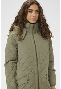 Zaļa dūraiņu jaka ar augstu apkakli, rāvējslēdzēju priekšpusē un spiedpogām aizdarāmām kabatām. Izceļas ar redzamu šuvju rakstu un mīkstu spīdumu vieglā materiālā.