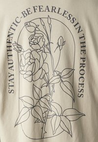 Beige stof met een zwart omlijnd ontwerp van een roos en bladeren. Tekst leest: "BLIJF ECHT - WEES VREESLOOS IN HET PROCES."