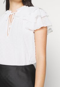 Kvinna i en vit, texturerad blus med korta, volangärmar och en knytdetalj vid halsen, i kombination med en svart kjol.