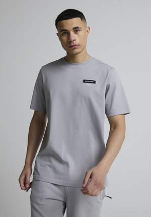 Ung mand med kort hår iført lysegrå t-shirt og matchende bukser, stående mod en ensfarvet grå baggrund.