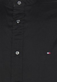 Camisa negra de algodón con botones. Presenta botones plateados y un pequeño logo en rojo, blanco y azul en el pecho izquierdo.