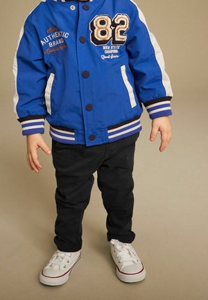 Veste bomber bleue avec des accents blancs, arborant un grand "82" brodé et des rayures sur les poignets, associée à un pantalon noir et des baskets blanches.