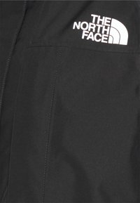 Czarna kurtka o gładkiej fakturze, z wyraźnym białym logo "THE NORTH FACE" na górnej lewej stronie.