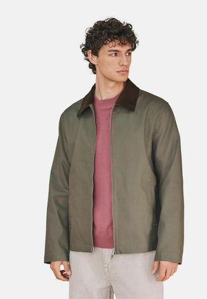 Junger Mann mit lockigem Haar, der eine olivgrüne Jacke über einem mauvefarbenen Pullover und hellbeige Hosen trägt und nach rechts schaut vor einfachem Hintergrund.