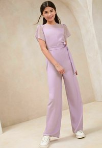 Lavendel-Jumpsuit mit transparenten kurzen Ärmeln, weitem Bein, Taillengürtel und glatter Textur. Getragen mit weißen Sneakers. Neutrale Hintergrund.