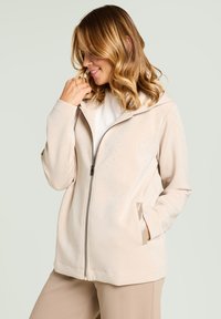 Veste beige zippée avec capuche, présentant de petites perforations, une texture lisse et des poches latérales. Conception légère et coupe décontractée.