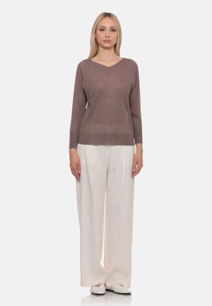 Donna in piedi che indossa un top marrone a maniche lunghe, pantaloni bianchi a gamba larga con coulisse e scarpe bianche su uno sfondo chiaro semplice.