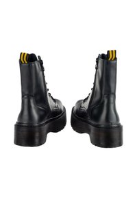 Botas de tobillo de cuero negro con cordones, suela gruesa, cremalleras verticales y una lengüeta de tira amarilla y negra en la parte trasera.