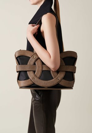 Black handbag featuring a textured brown strap design, with circular accents and silver hardware, held by a figure in a black top.