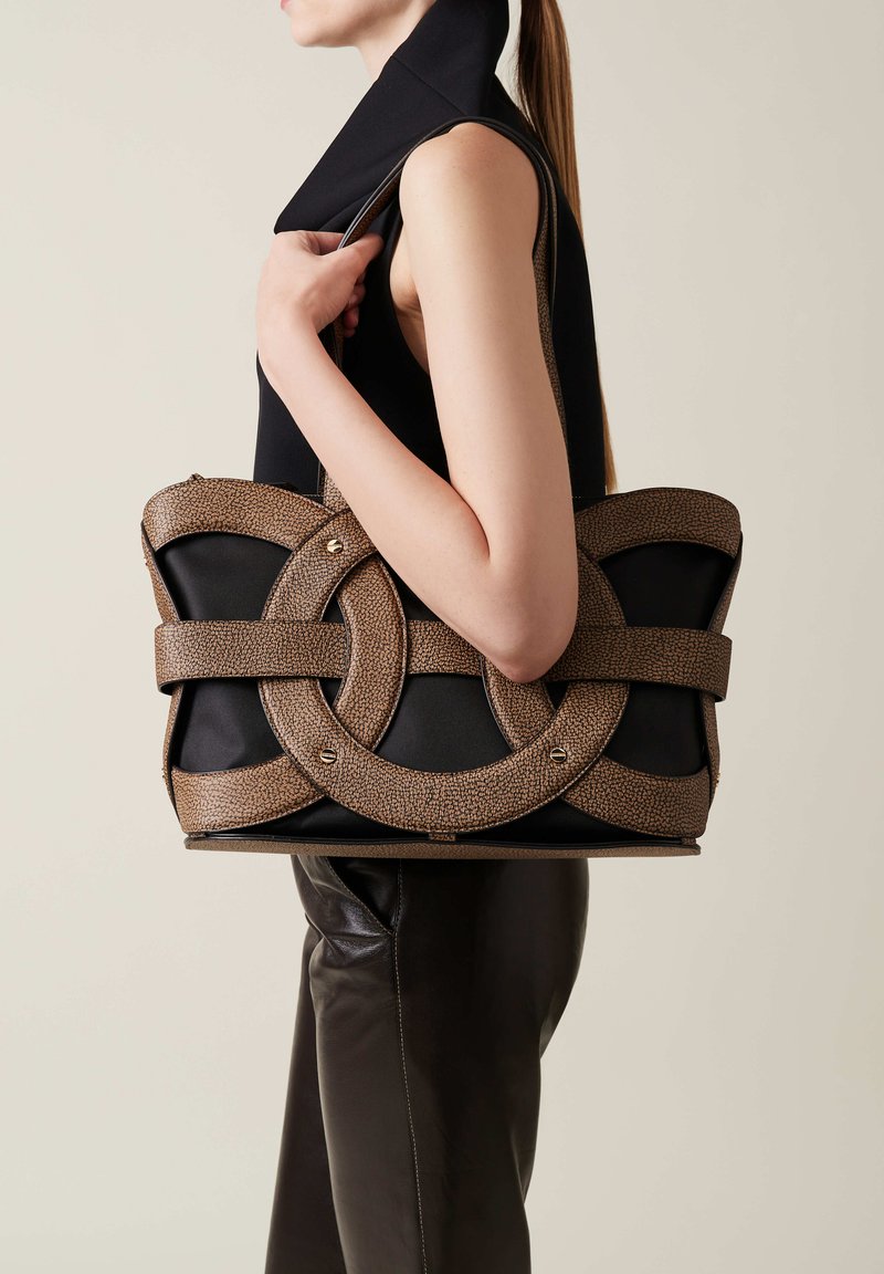 Sac noir avec une sangle texturée marron, ornée d'accents circulaires et de quincaillerie en argent, tenue par une personne en haut noir.