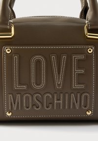 Καφέ τσάντα χειρός από συνθετικό δέρμα με ανάγλυφο κείμενο "LOVE MOSCHINO", χρυσά μεταλλικά στοιχεία και λευκό ράψιμο. Ορθογώνιο σχήμα με επίπεδη βάση.
