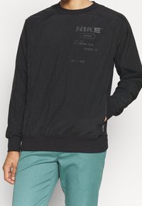 Sweatshirt noir à manches longues avec une finition texturée et un ourlet côtelé contrastant. Présente des éléments de marque imprimés et des détails dans une finition mate subtile.