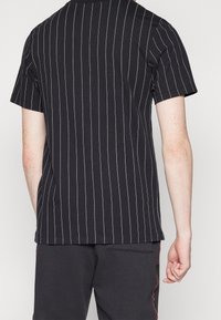 Černé tričko s bílými svislými pruhy, krátké rukávy a kulatým výstřihem. Materiál vypadá měkce; má volný střih a minimální detaily šití.