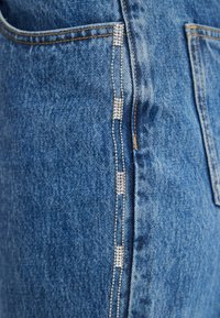 Siniset farkut, joissa pystysuora strassikoristelu sauman varrella, klassinen viiden taskun malli ja näkyvät saumaustekstit.