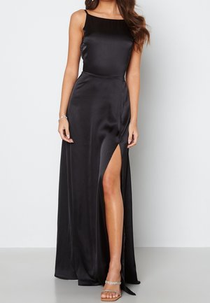 Femme portant une robe noire en satin sans manches avec une fente haute sur la cuisse et des sandales ornées de strass, debout sur un fond blanc.