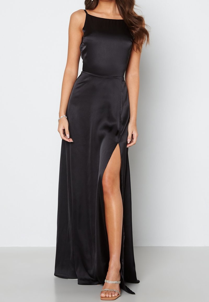 Femme portant une robe noire en satin sans manches avec une fente haute sur la cuisse et des sandales ornées de strass, debout sur un fond blanc.