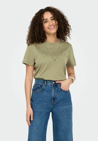 Oliven grøn T-shirt med korte ærmer og blomsterudskæringer, parret med højtaljede blå denim jeans. Glat stoftekstur.