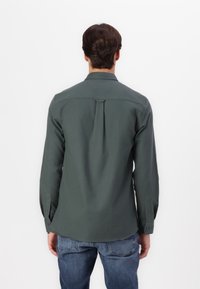 Cămașă cu mâneci lungi de culoare verde închis, cu tiv drept, manșete cu nasturi și un detaliu cu pliu pe spate. Fabricată dintr-un material neted cu un finisaj ușor texturat.