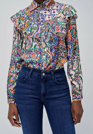 Gros plan d'une personne portant un chemisier à manches longues à motif paisley coloré avec un détail volanté, rentré dans un jean taille haute bleu foncé.