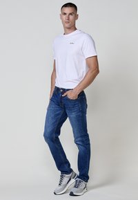 Camiseta de algodón blanca con un pequeño logo negro, combinada con jeans de mezclilla azul oscuro con costuras naranjas, y zapatillas grises con detalles en azul marino.